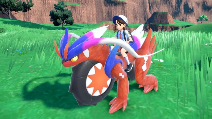 The Pokémon Koraidon. It is a red Pokémon with four legs and also large wheels, making it act like a motorbike. It is being ridden by a trainer.