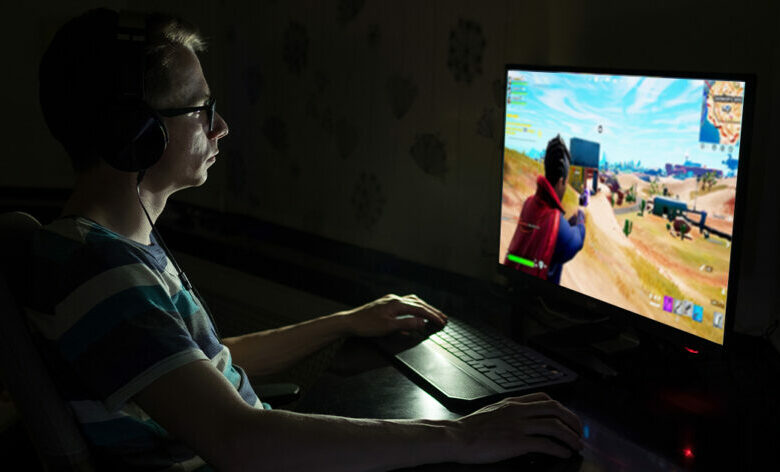 A man plays Fortnite in darkness on a PC computer.