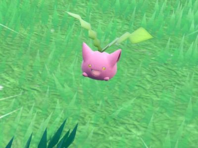 Hoppip, a round pink Pokémon with large ears and two green shoots growing from its head, against a grassy field.