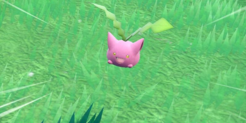 Hoppip, a round pink Pokémon with large ears and two green shoots growing from its head, against a grassy field.